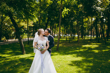 Beautiful wedding photosession. Handsome groom in blue formal suit and his elegant bride in dress and veil with bouquet with beautiful hairdress embrace on a walk in the big green park on sunny day