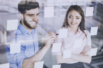 Fototapeta premium Young businessman and businesswoman working on new project with many sticky notes on glass screen in office