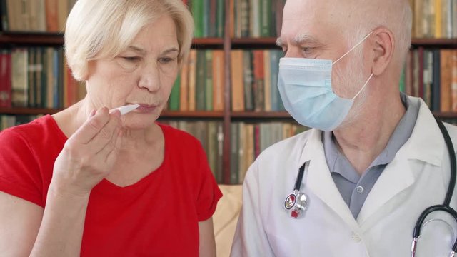 Male professional doctor in white coat and medical mask at work. Senior man physician measuring temperature to sick senior female patient by thermometer at home. Consulting about treatment and therapy