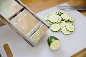 A pile of zucchini slices sits on a cutting board next to a mandolin