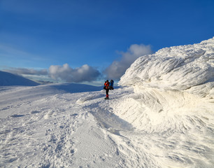 A panorama of snow capped mountains with textured, frozen, mystical fanciful shaped stones, a man in a ski suit with a backpack and a camera.