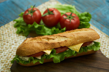Fresh submarine sandwiches with chicken breast cheese, tomatoes, lettuce, cucumbers and onions on dark wooden cutting board, rustic background