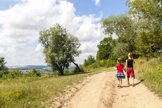 A boy and a girl children walk on a dirt road on a sunny summer day. Kids holding hands together while enjoying ativity outdoors.