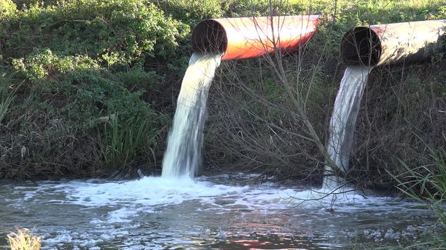 Contaminated water is dumped into a waterway through industrial pipes.