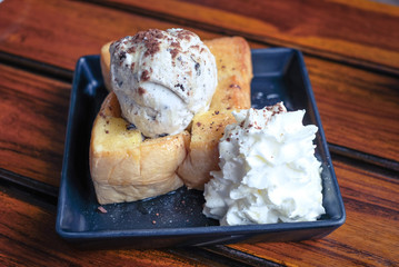 Honey toast with ice cream and whipped cream on black plate, wooden table.