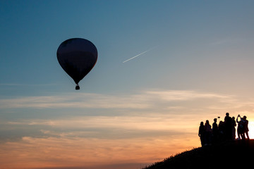 hot air balloon flies away into the sky and a lot of people watch after him on the top of the hill on the sunset