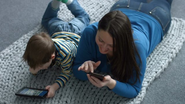 Charming single mother surfing the net on smart phone and her cute little boy playing on line game with digital tablet while lying on the floor at home. Top view. Happy family using technology devices