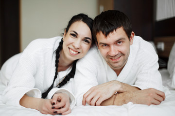 Fototapeta premium Young husband and wife of white coats play and smile in bed in a hotel room. Close up.