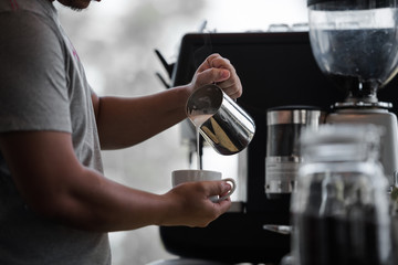 Hand of male barista holding and pouring milk for cup of coffee in the workplace at cafe