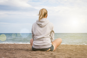 Fototapeta na wymiar Warm clear sunny summer day. A girl in a gray sweatshirt and jeans shorts sits with her back to the camera on the sad shore and looks at the sea