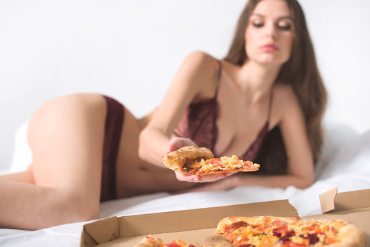 seductive woman lying in lace lingerie on bed and taking piece of pizza
