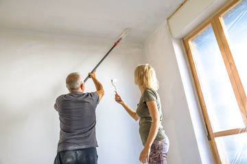 Caucasian housewife helping her whitewasher man at work painting a blank ceiling, with paint brush roller in empty white room for renovation. © bennymarty