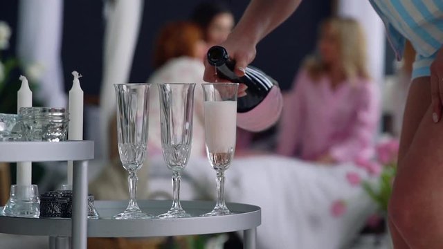 Close-up of a beautiful girl fills with champagne glasses for girlfriends who have fun in the background at a bachelorette party.
