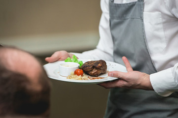 fresh grilled bbq roast beef steak and sauce on a white plate with green leaf of salad. soup sauce small jug glass served on a table in a restaurant