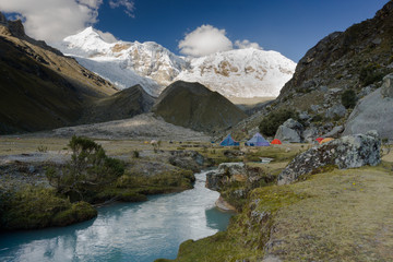 base camp in the Andes in Peru with snowy high peaks behind and large boulders and red rocks and a little mountain stream in the foreground