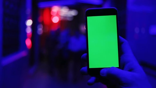 Man's hand using mobile smartphone with blank green screen in the nightclub / ultraviolet light blue walking people blur background chromakey tapping display swapping flapping tap touchscreen mockup