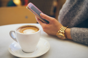 Close-up of white cup of coffee on the table in a modern cafe and woman hand with clock holding smartphone. Selective focus.
