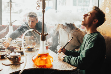 A young company of people is smoking a hookah and communicating in an oriental restaurant. Lebanon cuisine served in restaurant.  Traditional meze lunch - obrazy, fototapety, plakaty
