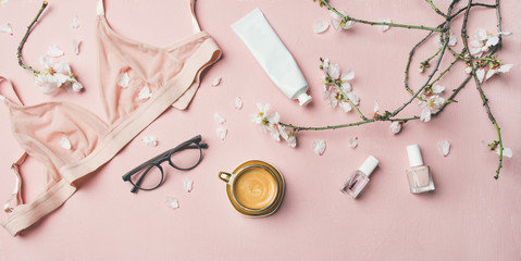 Womans morning rituals concept. Flat-lay of feminine tender powder color lingerie, glasses, cosmetic items, cup of coffee and Spring blossom flowers over pastel pink bed cover, top view.