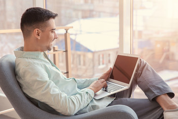 Young man with laptop sitting in armchair near window