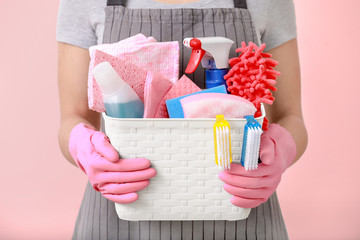 Woman holding basket with cleaning supplies on color background