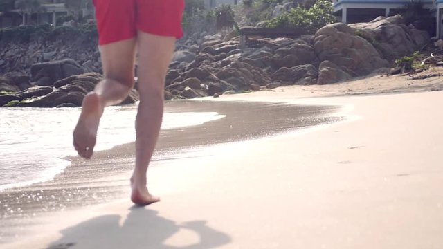 A slender man in red shorts runs along a sandy beach to large stones, sea waves roll to the shore. slow motion, 1920x1080, full hd