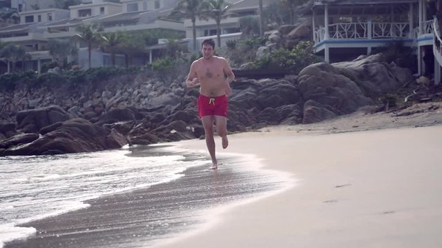 A slim man in red shorts runs along a sandy beach, sea waves roll to the shore. slow motion, 1920x1080, full hd