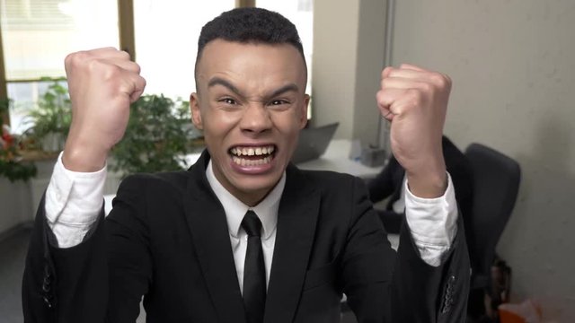 Young successful African businessman screams and raises his fists up, rejoices at the victory, looking at the camera, portrait, close-up. Man working at the computer in the background. 60 fps