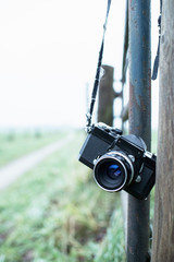 Lost vintage SLR camera hanging on fence post in countryside.