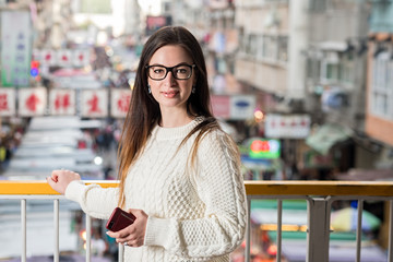 Young attractive caucasian brunette woman in eyeglasses standing on pedestrian footbridge and looking at camera. Blurred traditional asian street market as background. Urban portrait.