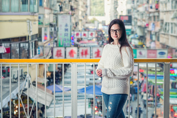 Young attractive caucasian brunette woman in eyeglasses standing on pedestrian footbridge and looking at camera. Blurred traditional asian street market as background. Urban portrait.