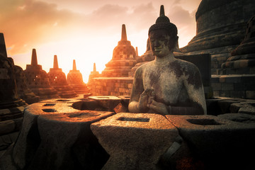 Amazing sunrise view of meditating Buddha statue and stone stupas against shining sun on background. Ancient Borobudur Buddhist temple. Great religious architecture. Magelang, Central Java, Indonesia