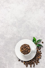 Coffee beans in white cup and green leaves on concrete background. Top view, space for text.
