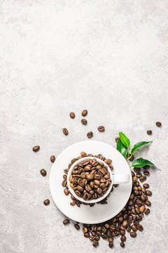 Coffee beans in white cup and green leaves on concrete background. Top view, space for text.