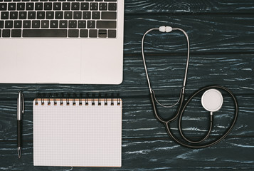 top view of arranged laptop, empty notebook and stethoscope on dark wooden tabletop