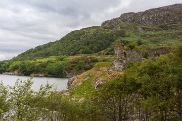Fototapeta na wymiar Stromeferry, Scotland - June 10, 2012: Castle Strome ruins on green shore cliff over Loch Carron. Cloudscape and forested mountain in back. Top of trees in front.