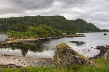 Stromeferry, Scotland - June 10, 2012: Loch Carron shoreline with peninsula under Castle Strome ruins. Green hills on shores. Motor boat anchored in lake at distance. Rain-heavy cloudscape.
