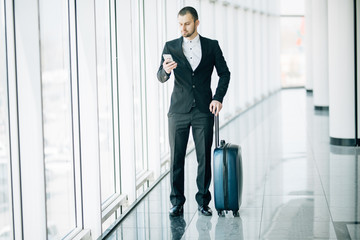 Elegant businessman checking e-mail on mobile phone while walking with suitcase inside airport terminal. Experienced male employer using cell telephone before work travel