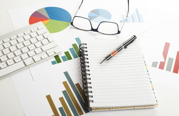 Background top view of workplace with economic graphics of various colors next to keyboard, pen and glasses with an open notebook in the middle of the image. Copy space. Mockup.