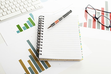 Background top view of workplace with economic graphics of various colors next to keyboard, pen and glasses with an open notebook in the middle of the image. Copy space. Mockup.