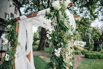 Wooden ceremony arch decoretade by white cloth, flowers and greenery standing in bright garden for wedding ceremony. Decor.