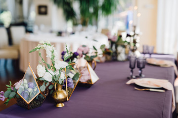 Composition of flowers in florariums standing on table covered by violet cloth and decorated by candles and cutlery. Decor. Wedding. Reception