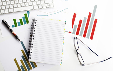 Background top view of workplace with economic graphics of various colors next to keyboard, pen and glasses with an open notebook in the middle of the image. Copy space. Mockup.