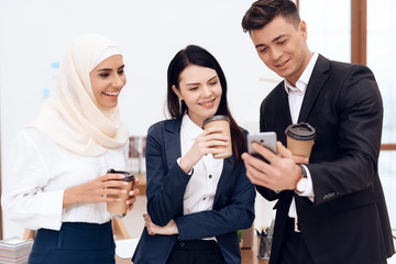 Two women and a man are drinking coffee and watching something on the phone screen.