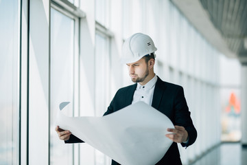 Portrait of a smiling male contractor engineer with hard hat reading blueprint at construction site.