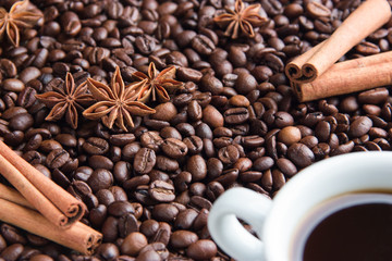 a cup of espresso coffee on the right in the corner on roasted coffee beans spilled on a table with cinnamon sticks and anise