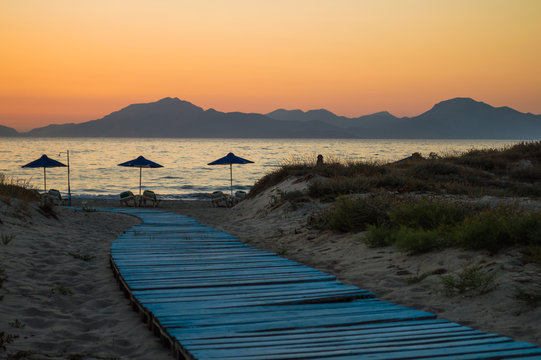 A light blue boardwalk leads down to the umbrellas on the beach, as the sun set behind the mountains at golden hour.