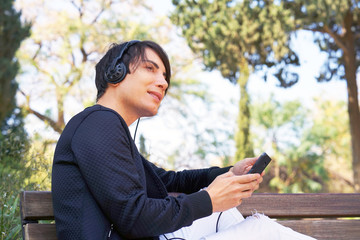 Fototapeta premium The guy gay is sitting on the bench, using a smartphone to listen to music on headphones. Joyful attractive gay listening to music on headphones while enjoying sitting in the park. Lifestyle concept