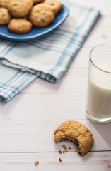 A glass of milk and a cookie with a bite taken out of it next to a blue plaid napkin with a plate of cookies in the background