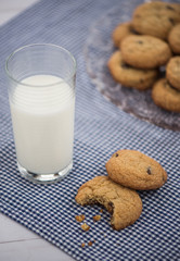 A glass of milk and a cookie with a bite taken out of it on a blue gingham napkin with a plate of cookies in the background